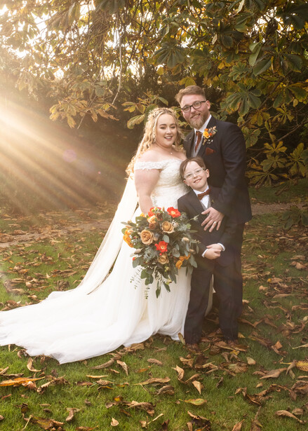 Bride and groom posing outdoors with a young child, golden sunlight filtering through trees behind them.