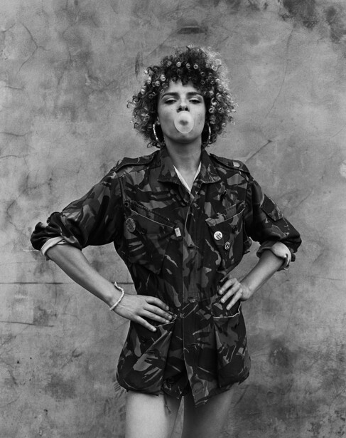 Person with voluminous curly hair wearing a black leather jacket against a textured gray background.