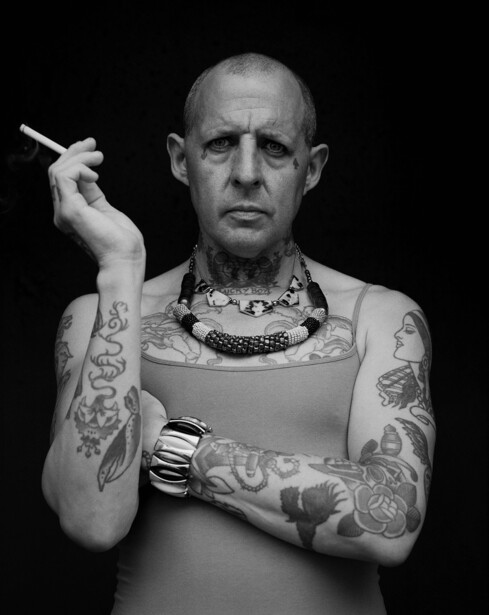 Shirtless man with extensive tattoos holding a cigarette, photographed against a black background.