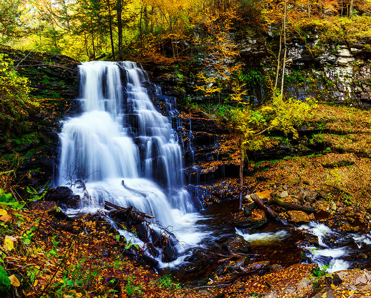 Multi-tiered waterfall cascading over dark rocks surrounded by golden autumn foliage.