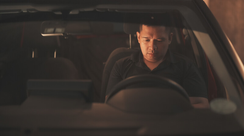 Man sitting in driver's seat of car at night, hands on steering wheel, looking ahead with serious expression.