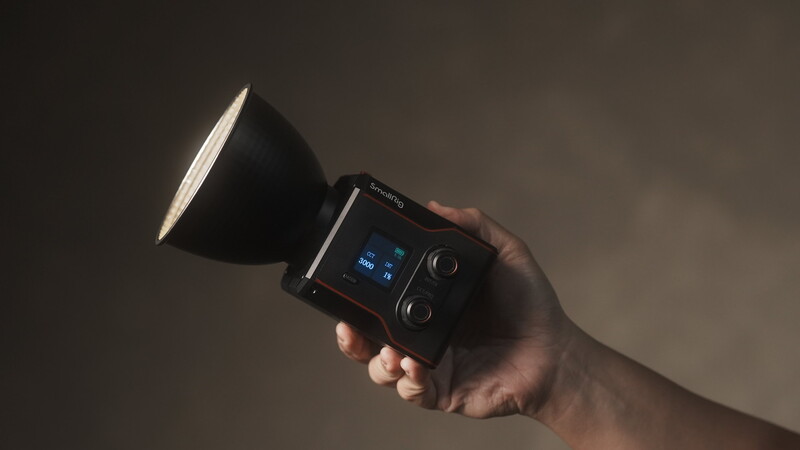 Hand holding a portable flash unit with a small LCD screen and control dial against a neutral gray background.