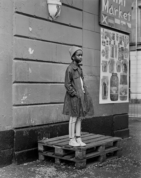 Young girl standing on wooden pallets against a building wall with storefront signage.