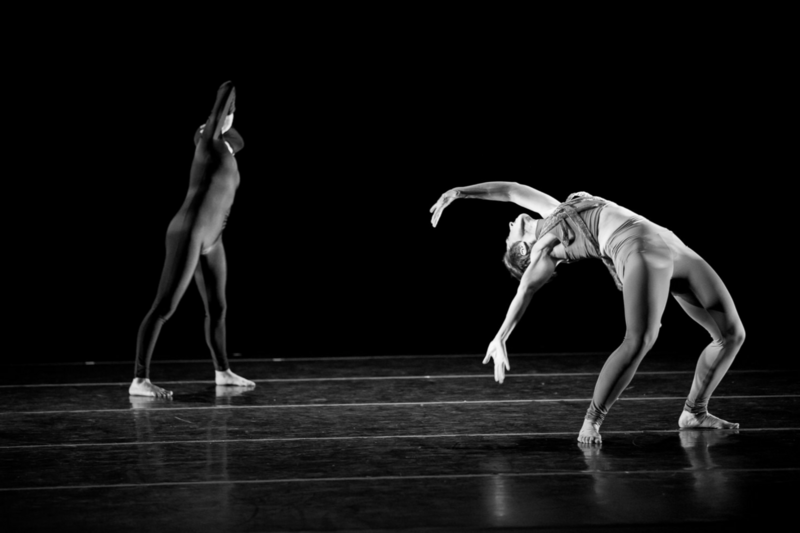 Two dancers in contrasting poses on a dark stage, one standing upright with arm raised, the other bending forward with curved spine.