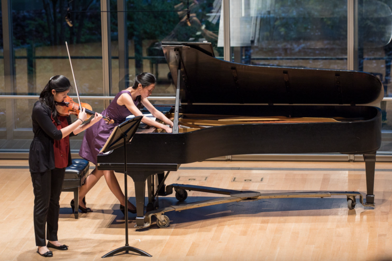 Two musicians perform at a grand piano during a classical concert in a modern venue with large windows.