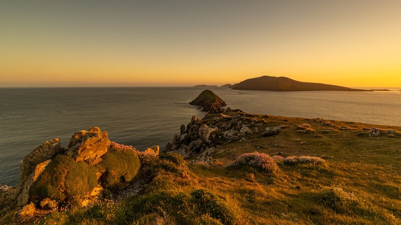 Rocky coastline at golden hour with distant islands silhouetted against a warm sky.