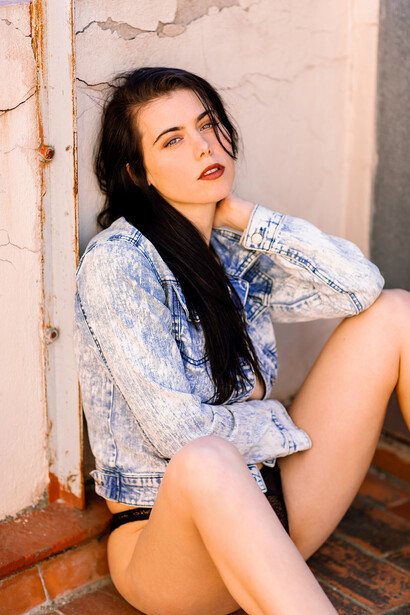 Woman with long dark hair sitting against a weathered white wall, wearing a light blue patterned shirt.