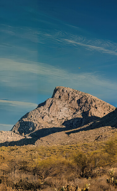 Rocky desert mountain peak rising above sparse scrubland under blue sky with wispy clouds.