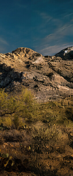 Rocky hillside with sparse desert vegetation under blue sky with wispy clouds.