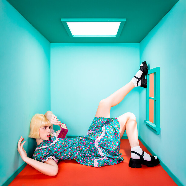 Woman lying on red floor in bright turquoise room, wearing patterned dress and black shoes.