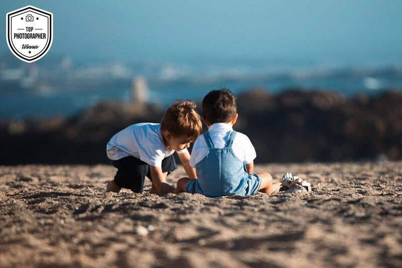 Two young children playing in sand on a beach, with a blurred cityscape and water in the background.