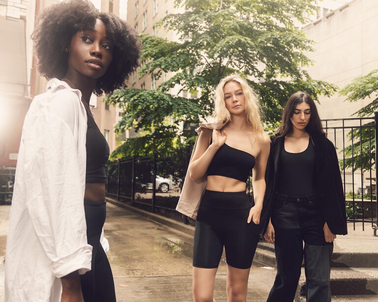Three women posing together on a wet urban street lined with ivy-covered buildings.
