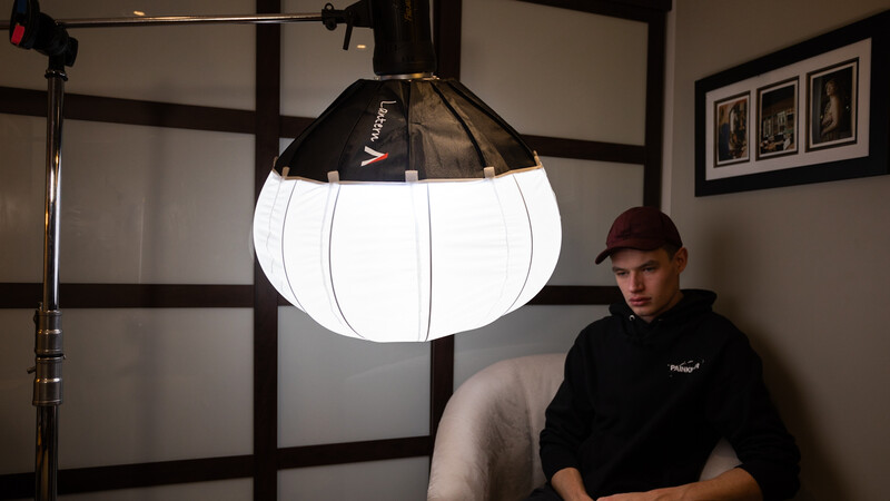 Large fabric softbox with vertical ribbed panels mounted on studio stand next to seated photographer.