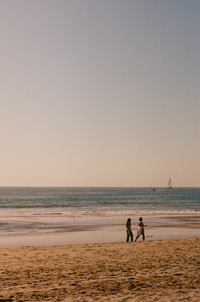 Two figures walking along a beach at golden hour with a sailboat visible on the horizon.