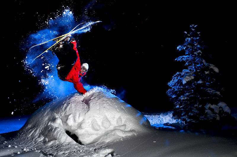 Athlete in red jacket jumping off snowy mound with blue light trails and winter trees.