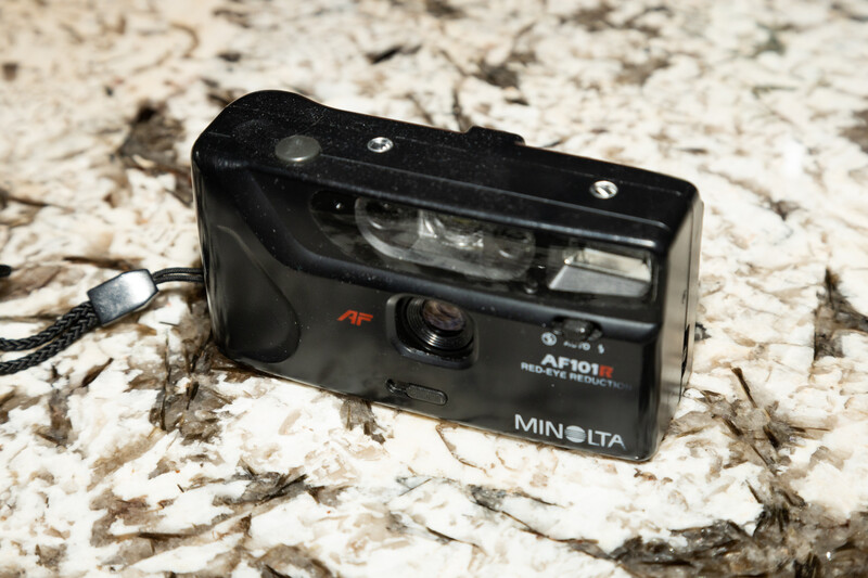 Minolta compact film camera with flash attachment on textured stone surface.