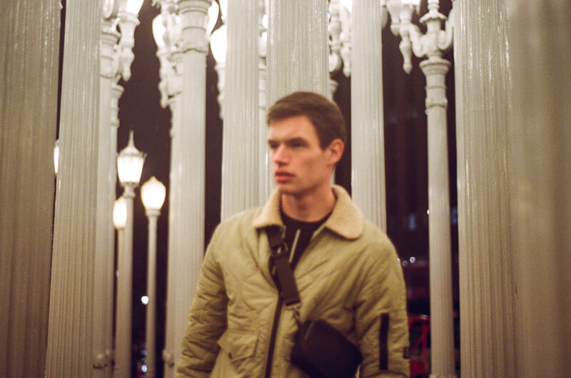 Man in tan jacket standing in modern corridor with illuminated architectural columns.