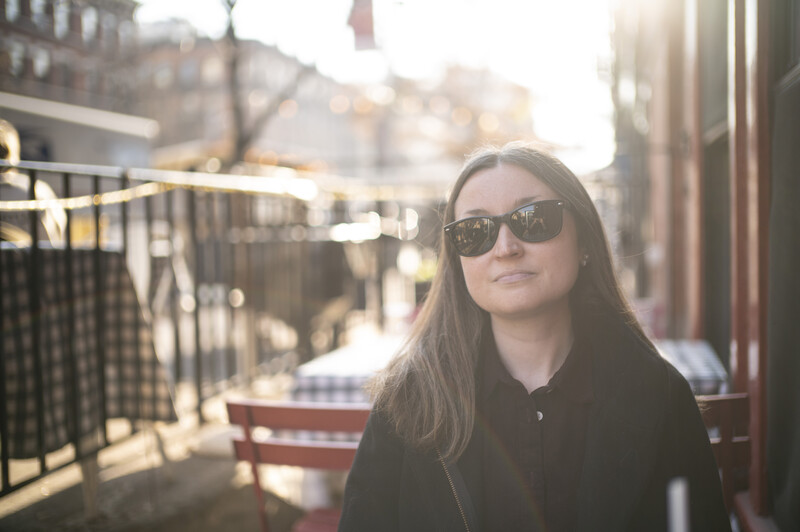 Woman wearing sunglasses and dark jacket photographed outdoors in golden hour light.