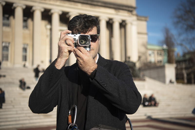 Photographer holding a vintage rangefinder camera up to his face in front of a classical building with columns.