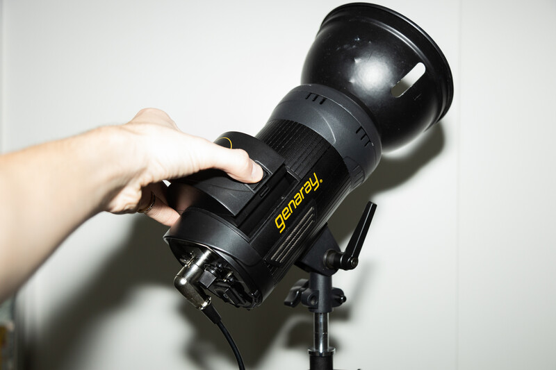Hand holding a Godox studio flash unit mounted on a light stand against a white wall.