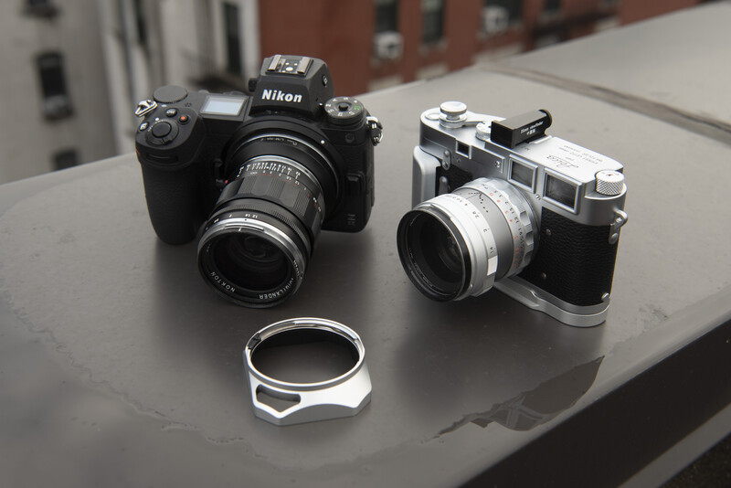 A modern Nikon DSLR camera next to a vintage rangefinder camera and a lens hood on a gray surface.