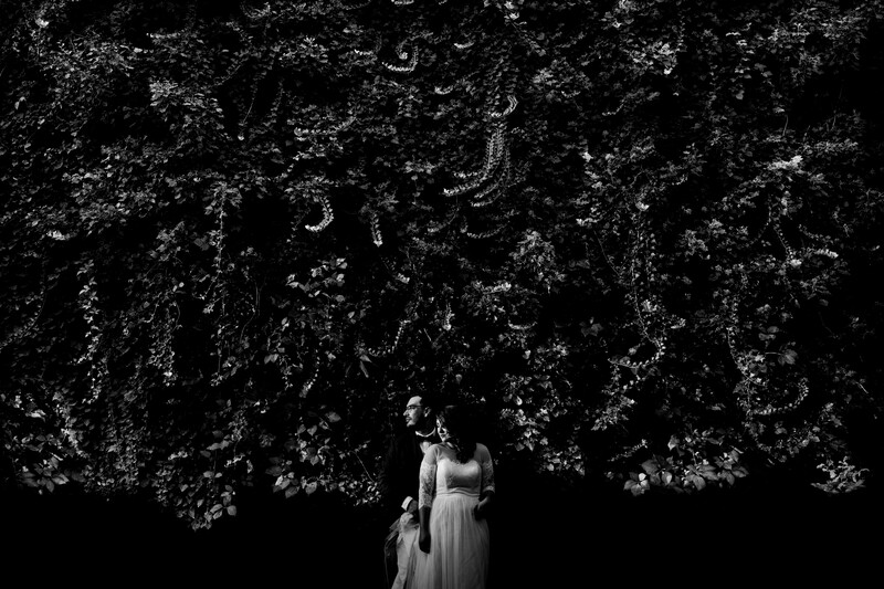 Woman in white dress standing beneath dense foliage canopy in dramatic black and white portrait.