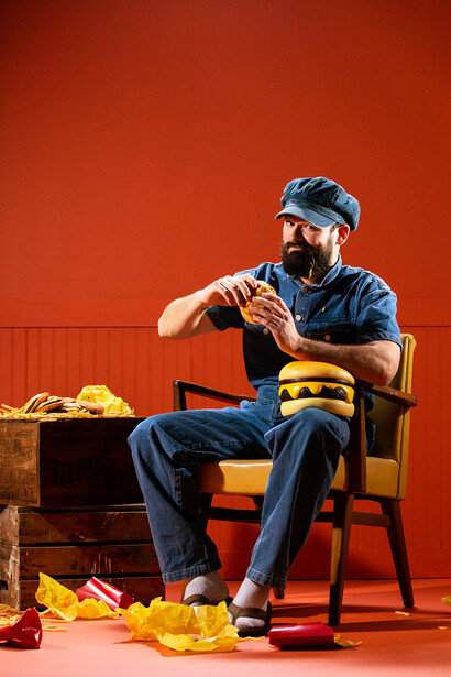 Man in denim shirt and cap sitting on wooden chair against vibrant red background, gesturing while speaking.