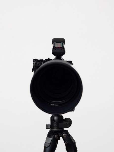 Large telephoto lens mounted on tripod with flash hot shoe adapter and external flash, photographed against neutral background.