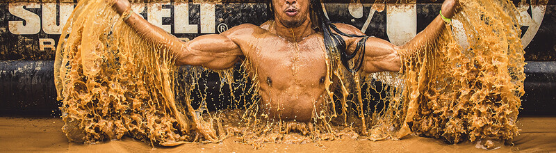 Shirtless athlete emerging from muddy water with arms spread wide, holding dripping fabric.