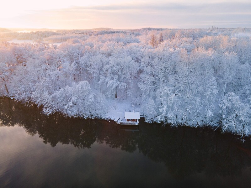 Frost-covered forest landscape with a small white house beside a dark river at sunrise.