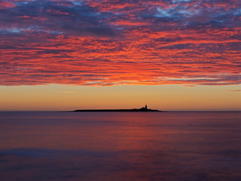 Silhouetted island with lighthouse against dramatic red and orange sunset sky reflected in calm water.