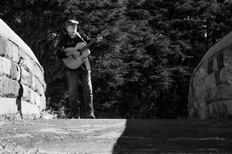 Musician with acoustic guitar standing between two concrete structures in a wooded area.