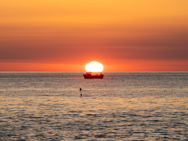 Large sun touching the ocean horizon at sunset, with a lone figure wading in shallow water.