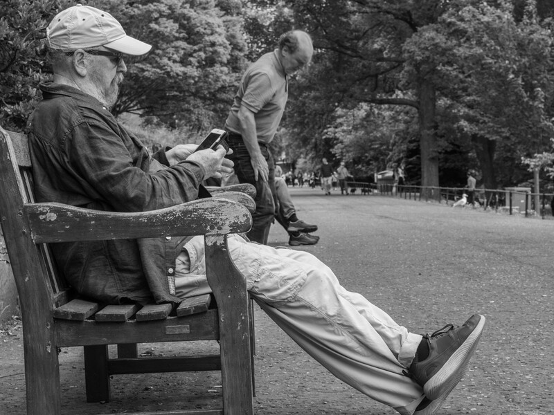 Man seated on a park bench reviewing photographs on a mobile device while another person stands nearby.