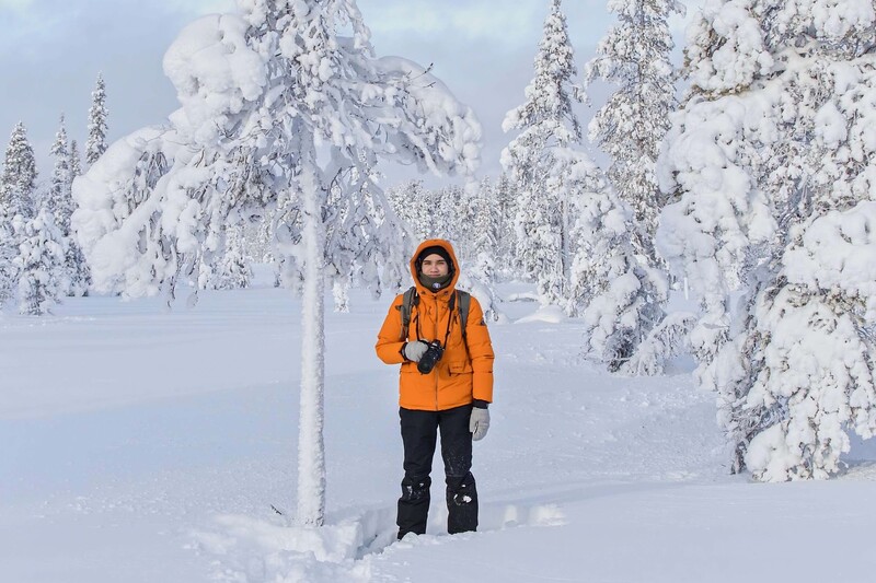Person in bright orange jacket standing in deep snow surrounded by frost-covered evergreen trees.