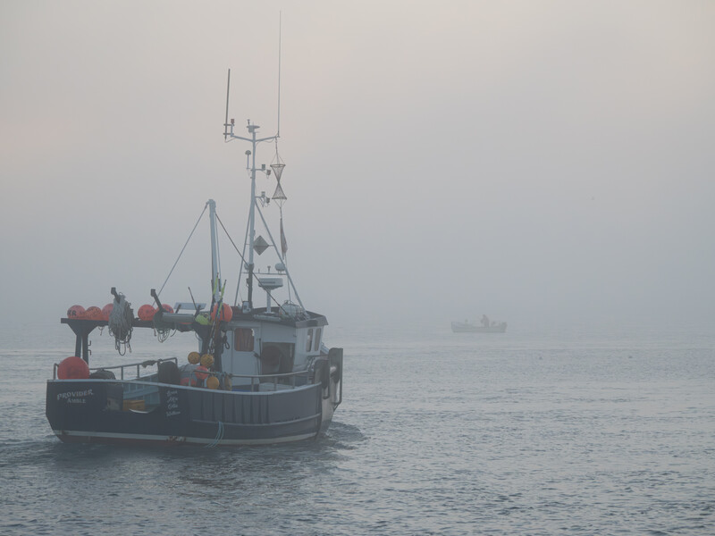 Commercial fishing vessel in heavy fog with antenna and rigging visible, another boat faintly seen in background.