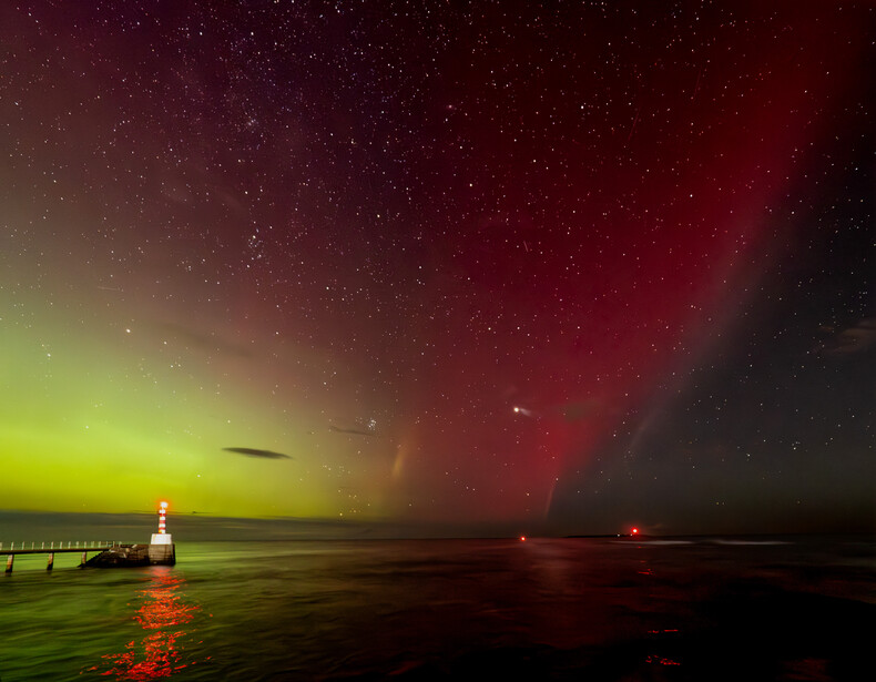 Aurora borealis illuminates the night sky above a coastal landscape with a lighthouse and pier.