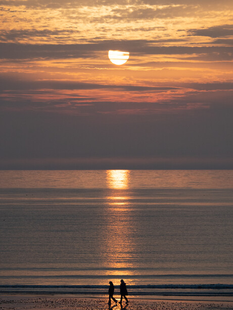 Two silhouetted figures walking along a beach during golden hour sunset with calm ocean water.