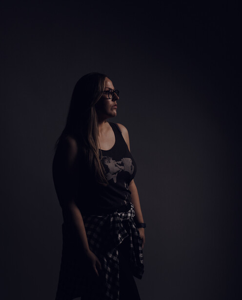 Woman in dark clothing with long dark hair standing against a black background, looking to the side.
