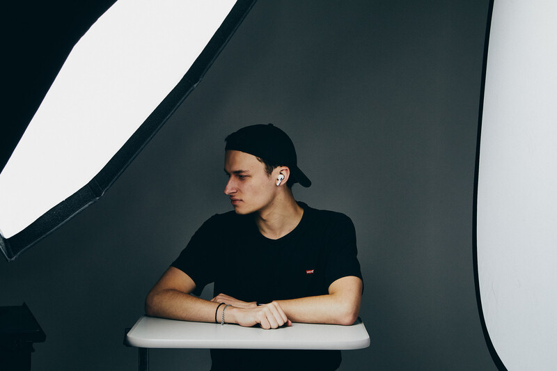 Man in black t-shirt and cap leaning against white surface in studio setting.