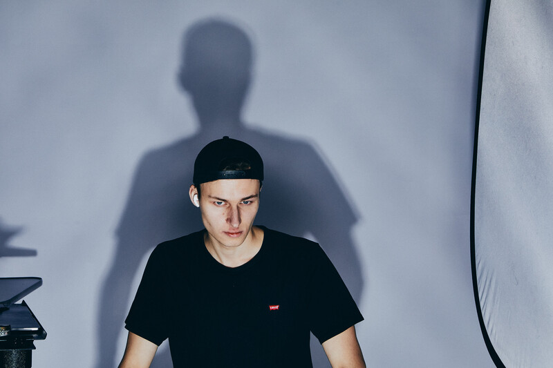 Portrait of a young man in a black t-shirt and cap against a white wall, with his shadow cast prominently behind him.