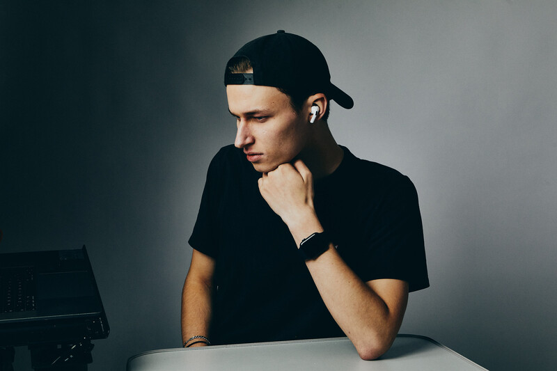 Man in black cap and t-shirt sitting at desk with thoughtful expression, hand to chin.