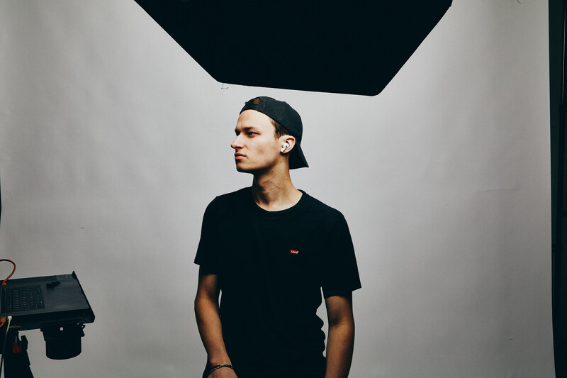 Young man in black t-shirt gazes to the side against minimalist white wall with dark geometric shape overhead.