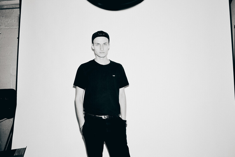 Man in black t-shirt and pants standing against white backdrop under studio lighting.