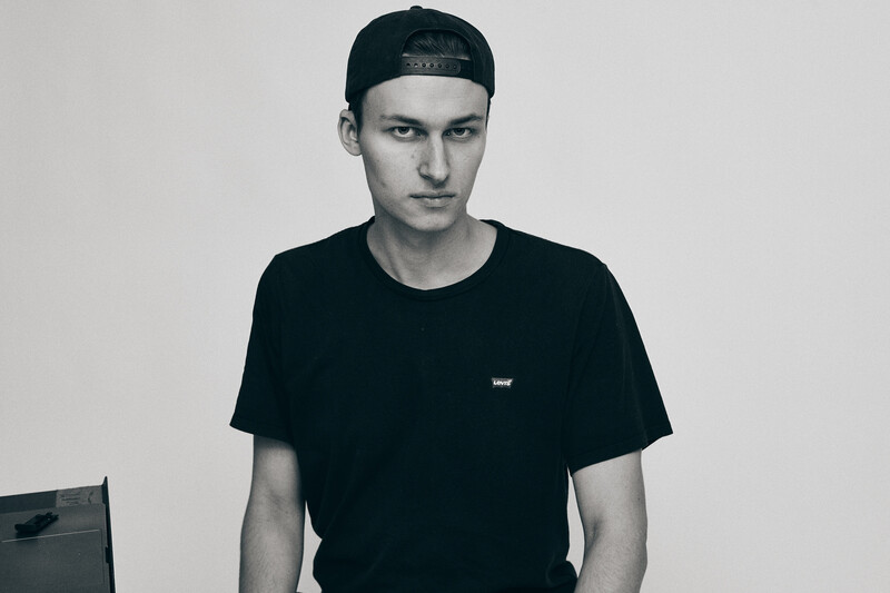 Young man wearing black t-shirt and cap looking directly at camera against plain white background.