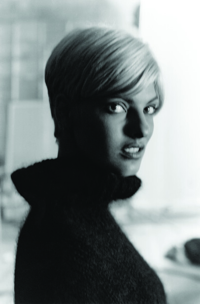 Black and white portrait of a woman with short blonde hair, shot in three-quarter view against a blurred interior background.