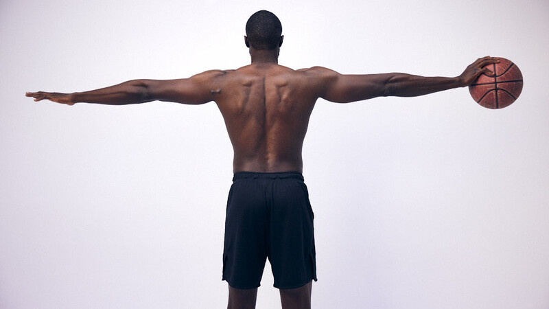 Shirtless male athlete viewed from behind with arms extended wide, holding a basketball in his right hand.