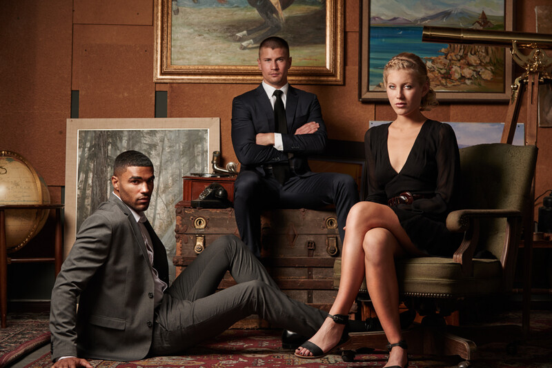 Three professionals in business attire posed in a vintage-styled office with globe, paintings, and leather furniture.