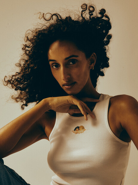 Woman with voluminous curly dark hair wearing a cream ribbed sleeveless top, posed with hand near face against neutral beige background.