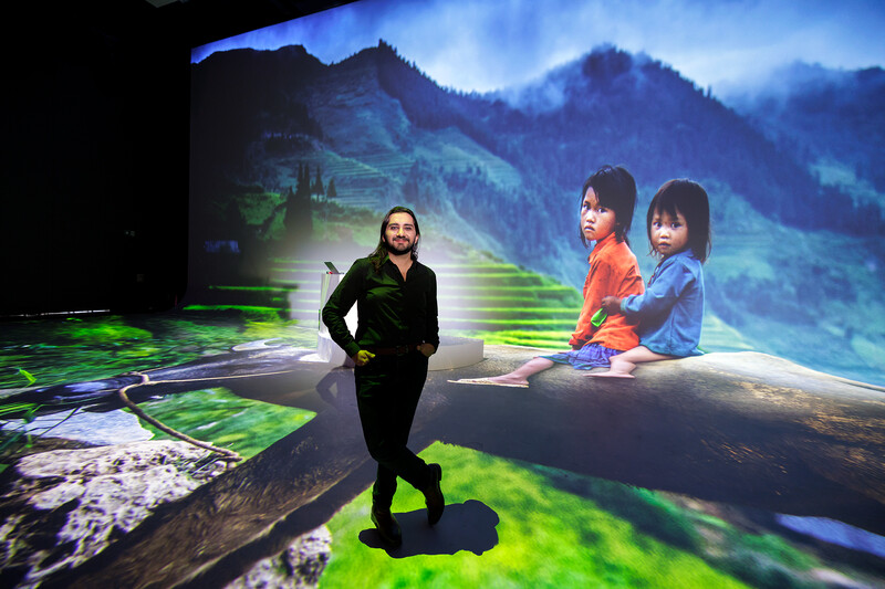 Man in black jacket standing on a log with mountainous landscape and two children projected behind him.